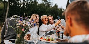 Kaksi naista ja mies istuvat vierekkäin ulkona puutarhassa ja ottavat selfie-kuvaa itsestään