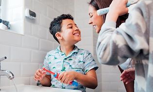 Poika esittelee äidilleen hampaita kylpyhuoneessa ja toinen lapsi harjaa hampaitaan