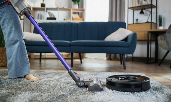 Ihminen imuroi mattoa varsi-imurilla ja vieressä on robottipölynimuri