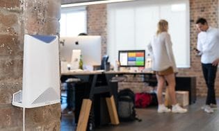 Netgear Orbi -mesh-reititin kiinnitettynä toimiston seinälle jossa kaksi työntekijää seisoo työpöydän vieressä juttelemassa