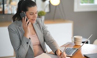Toimistossa istuva nainen puhuu puhelimeen ja tekee samalla muistiinpanoja