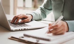 Nainen työskentelee kannettavalla tietokoneella ja kirjoittaa muistiinpanoja lehtiöön