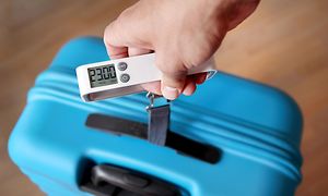 Käsi mittaamassa sinisen matkalaukun painoa matkatavaravaa'an avulla