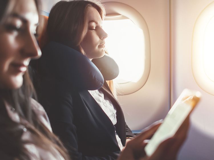 Kaksi lentokoneessa olevaa naista rentoutuvat, toisella on matkatyyny ja toinen katsoo puhelintaan