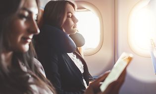 Kaksi lentokoneessa olevaa naista rentoutuvat, toisella on matkatyyny ja toinen katsoo puhelintaan