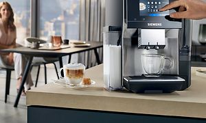 Siemens EQ700 -espressokone ja kupillinen kahvia keittiön työtasolla sekä taustalla istuva nainen