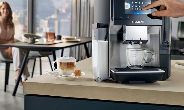 Siemens EQ700 -espressokone ja kupillinen kahvia keittiön työtasolla sekä taustalla istuva nainen