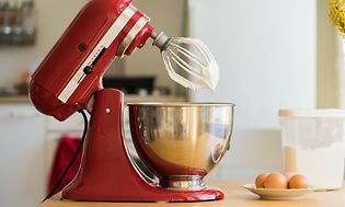 SDA - Pöydällä oleva KitchenAid-yleiskone sekä kananmunia ja rasiallinen hienoa sokeria sen vieressä