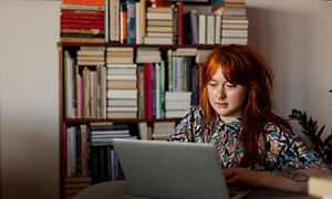 Nainen istuu macbook-kannettavan äärellä kirjahyllyn edessä