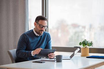 Liikemies istuu työpöydän ääressä kannettava vierellään ja käyttää tablettia