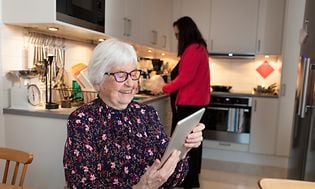 Hymyilevä iäkäs nainen istuu ruokapöydän ääressä ja käyttää tablettia