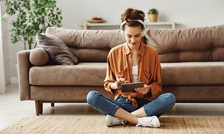 Nainen istuu sohvan edessä olohuoneen lattialla ja käyttää tablettia
