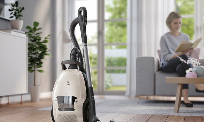 Electrolux-pölynimuri olohuoneen lattialla, jonka vieressä nainen istuu sohvalla kirjaa lukemassa