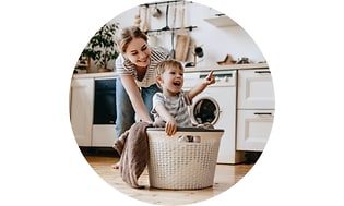 Keittiössä oleva äiti ja lapsi leikkivät pyykkikorin ympärillä