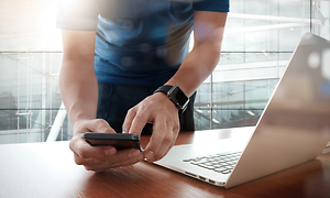 B2B - Android Zero-Touch - A man with his phone in front of his laptop in an office