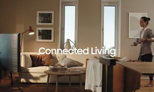 Nainen kävelee olohuoneeseen Samsung-puhelin ja kahvikuppi kädessään ja kuvassa teksti Connected living