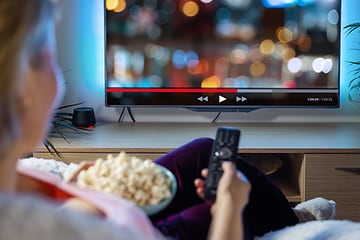 Nainen katselee televisiota ja pitelee kaukosäädintä kädessään ja popcorn-astiaa toisessa kädessään