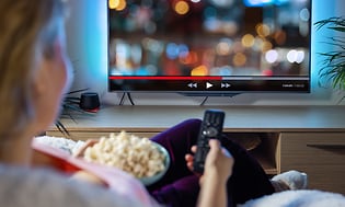 Nainen katselee televisiota ja pitelee kaukosäädintä kädessään ja popcorn-astiaa toisessa kädessään