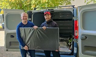 Kaksi miestä hymyilee kameralle kantaessaan televisiota pakettiautosta