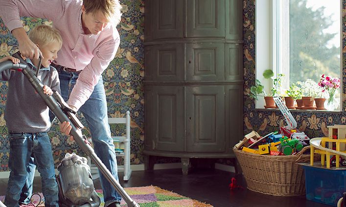 Isä auttaa pientä lastaan imuroimaan hänen huoneensa värikästä mattoa