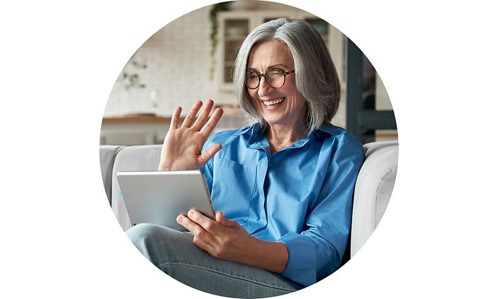 Vanhempi nainen istuu sohvalla ja vilkuttaa tabletin kameralle