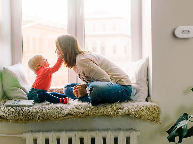 Seinällä oleva Airthings-ilmanlaatumittari sekä äiti ja lapsi yhdessä hempeilemässä