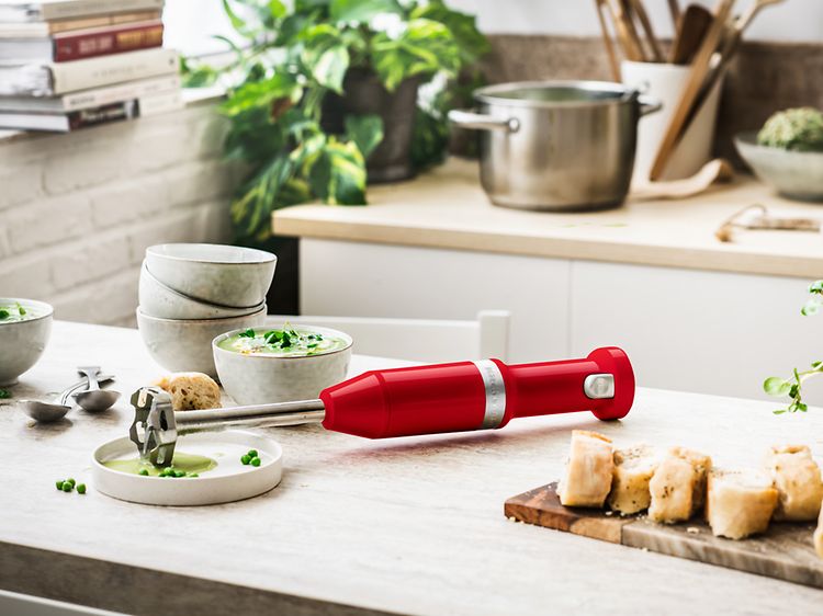 Punainen Kitchen Aid -sauvasekoitin keittiön työtasolla, jonka vieressä kulhollinen keittoa ja viipaloitua leipää