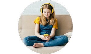 Koululaistyttö puhelin käsissä hymyilee ja istuu sohvalla kuulokkeet päässä
