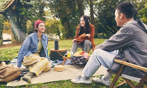 Kaksi naista ja mies istuvat puistossa piknikillä langaton kaiutin heidän keskellään korin päällä