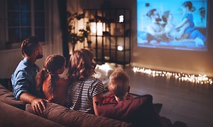 Perhe istuu sohvalla katselemassa elokuvaa projektorilla, joka heijastaa kuvan valkokankaalle