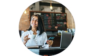 Kahvilassa istuskeleva nainen kannettava tietokone edessään ja matkapuhelin kädessään