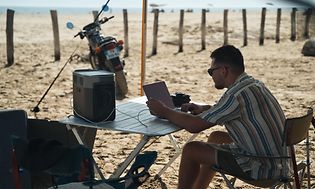 Mies istuu rannalla pöydän ääressä ja työskentelee kannettavalla, joka latautuu virta-aseman avulla