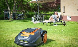 Nainen rentoutuu puutarhassa samalla, kun robottiruohonleikkuri siistii nurmikkoa