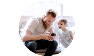 Isä istuu pienen tyttärensä kanssa sohvalla ja katsovat isän pitelemää puhelinta