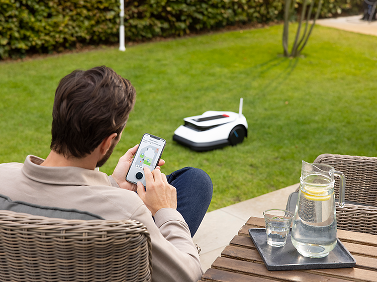 Ecovacs GOAT robottiruohonleikkuri leikkaamassa nurmikkoa puutarhassa ja mies ohjaa sitä älypuhelimellaan