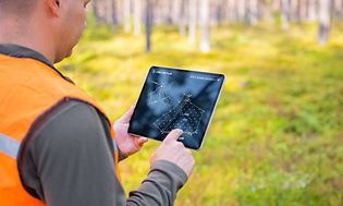 Oranssiin heijastinliiviin pukeutunut mies tarkastelee metsän aluesuunnitelmaa tabletilta