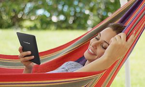 Ulkona riippumatossa makaava nainen katsoo Pocketbook-e-kirjanlukijaansa hymyillen