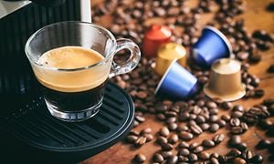 Kupillinen mustaa kahvia ja erilaisia kahvikapseleita pöydälle levitettyjen kahvipapujen päällä
