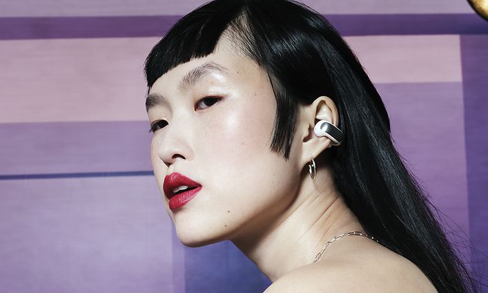 Avoimet Bose Ultra -nappikuulokkeet - violetin taustan edessä olevalla naisella on korvissaan open-ear-nappikuulokkeet