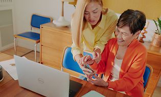 Kaksi naista käyttämässä kannettavaa tietokonetta ja älypuhelinta yhdessä