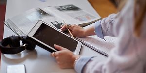Nainen pitelee tablettia oikeassa kädessään ja tarkastaa tietoja pöydällä olevasta kirjasta