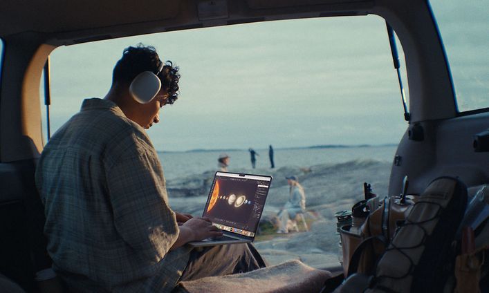 Nuori mies opiskelee rantamaisemissa MacBook Air -läppärillä Airpods Max -kuulokkeet korvissaan