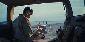 Nuori mies opiskelee rantamaisemissa MacBook Air -läppärillä Airpods Max -kuulokkeet korvissaan