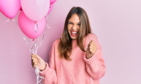 Iloisesti hymyilevä vaaleanpunaiseen paitaan pukeutunut nainen pitelee pinkkejä ilmapalloja kädessään