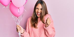 Iloisesti hymyilevä vaaleanpunaiseen paitaan pukeutunut nainen pitelee pinkkejä ilmapalloja kädessään