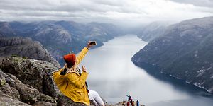 Keltaiseen takkiin pukeutunut nainen istuu kallion reunalla ottamassa selfietä puhelimellaan