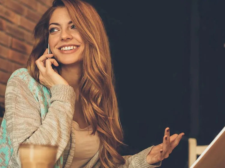 hymyilevä nainen kannettavan tietokoneen ääressä puhumassa puhelimeen