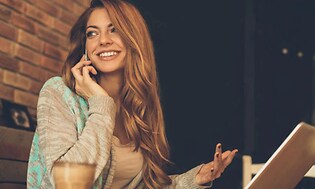 hymyilevä nainen kannettavan tietokoneen ääressä puhumassa puhelimeen