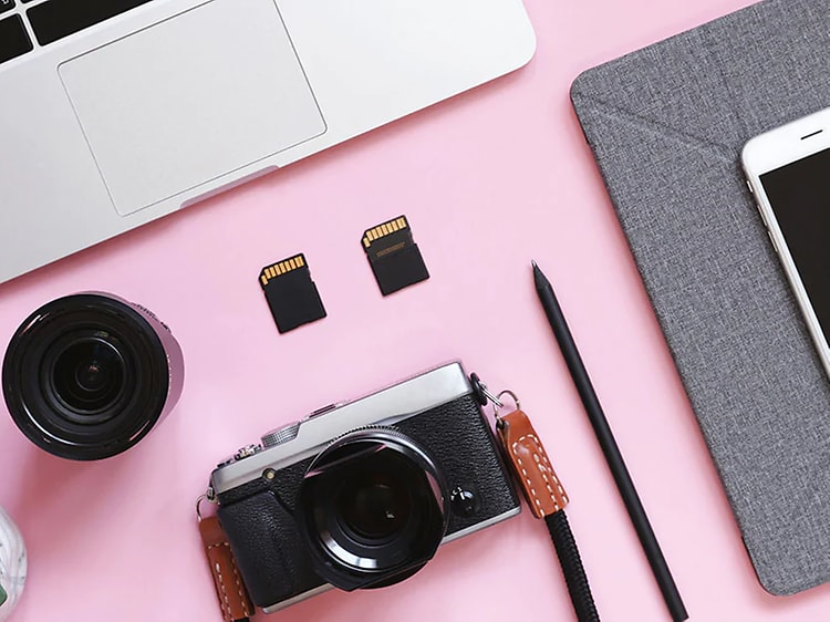 Valokuvaustarvikkeita, MacBook ja iPhone vaaleanpunaisella pöydällä