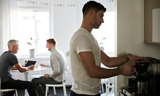 Toimistotyöntekijä kahvinkeitossa toimistossa, taustalla istuu kaksi kolleegaa pöydän ääressä